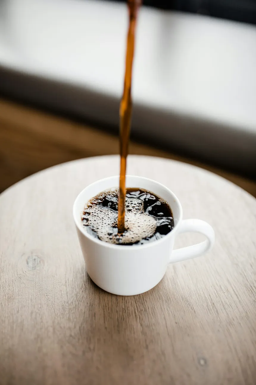 A close-up shot of black coffee being poured into a white mug on a wooden surface.