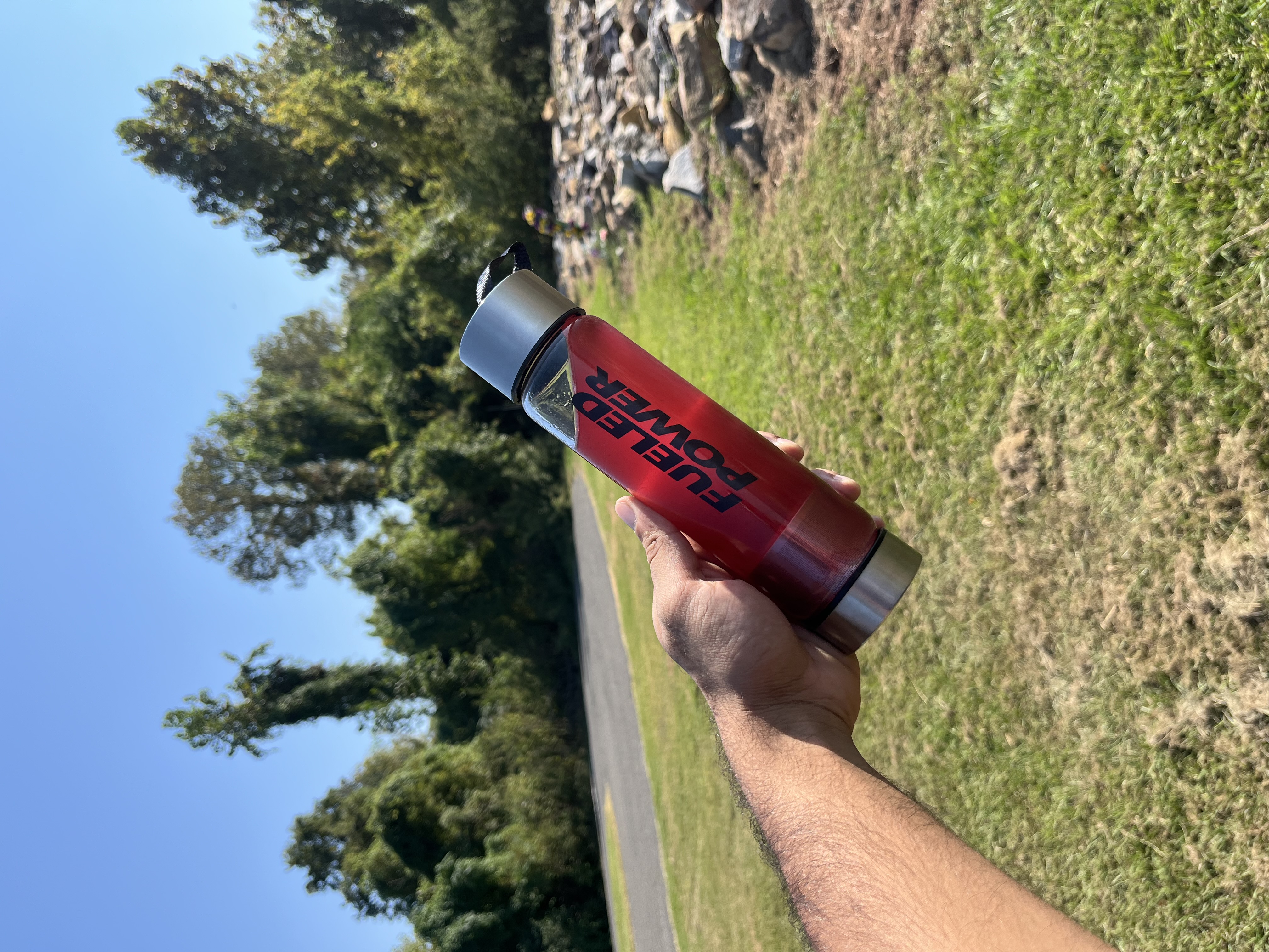 A person's hand holds a water bottle with red liquid outdoors in a grassy park on a sunny day.