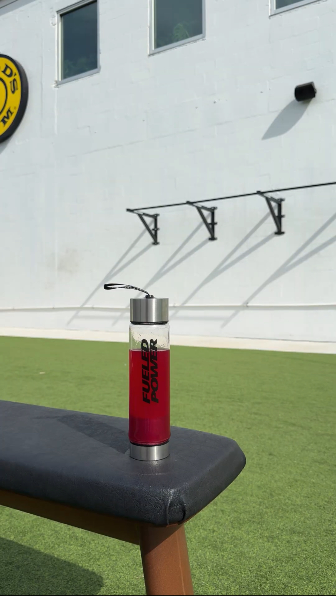 A glass bottle with red liquid and the words 'Fueled Power' sits on a workout bench outdoors.