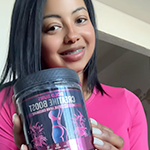 A smiling woman in a pink shirt holds up a black jar of creatine supplement.