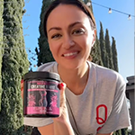 A smiling woman in a grey t-shirt holds up a black and pink jar of supplements outdoors.