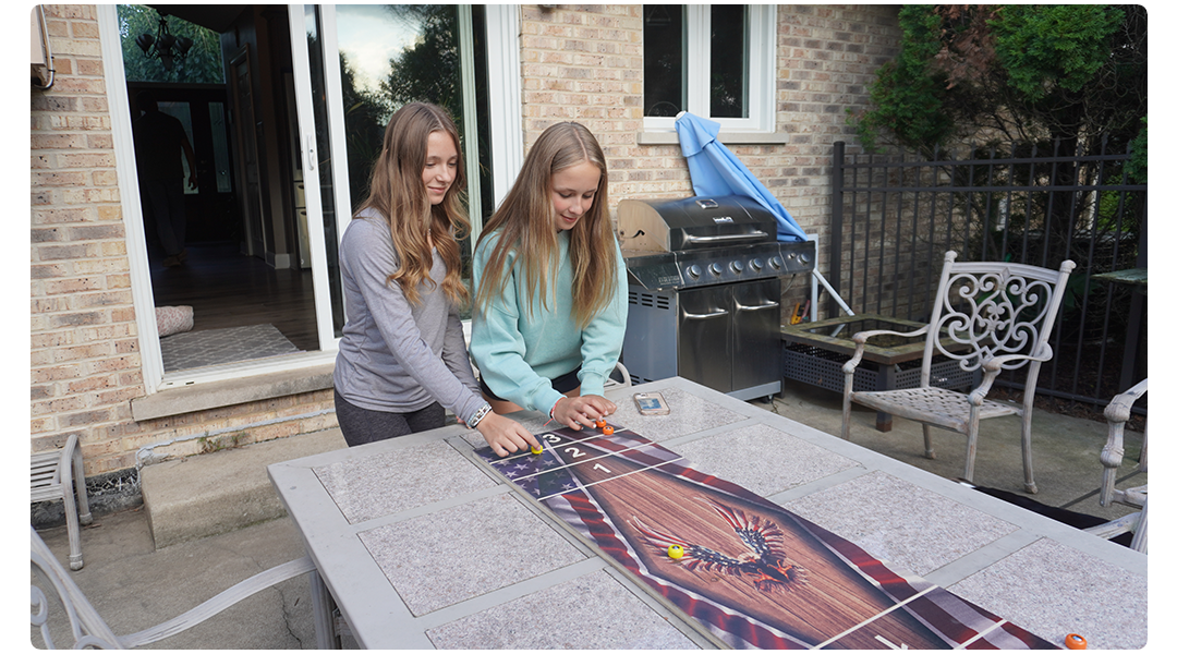 Two people playing a tabletop shuffleboard game outdoors near a barbecue grill.