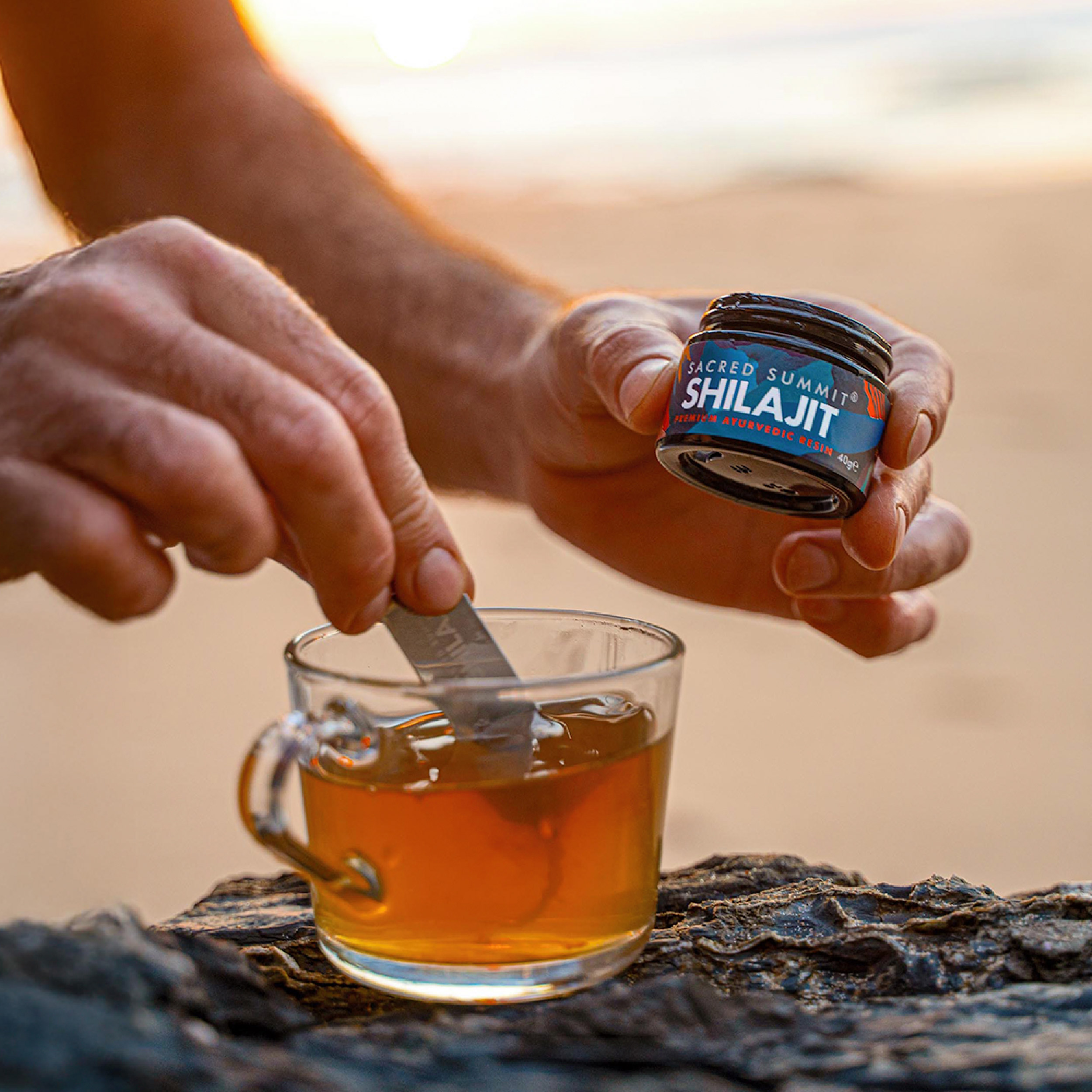 Person adding Shilajit to a cup of tea on a rocky surface.