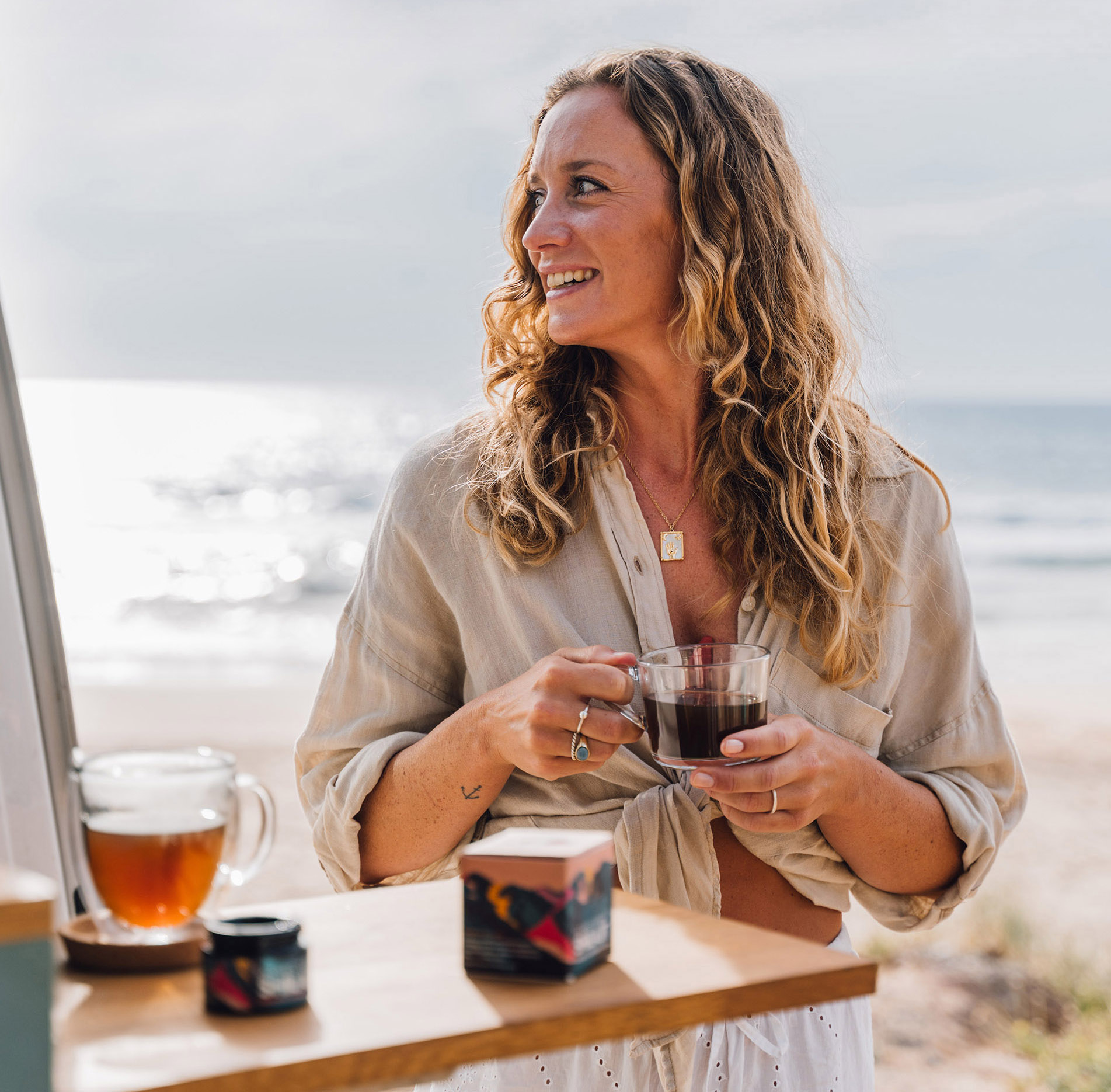 Woman smiling, holding a glass cup, standing by the beach.