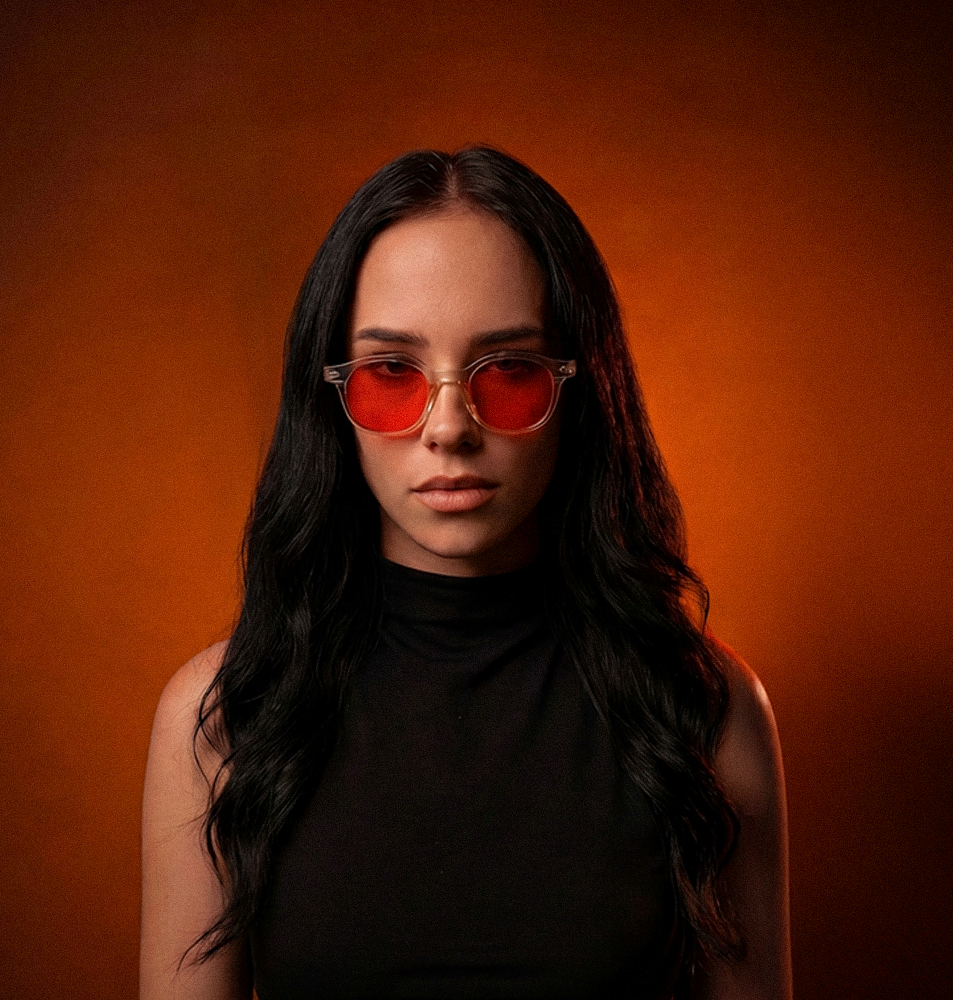 A woman with long black hair, wearing red-lensed glasses, looks at the camera against an orange background.