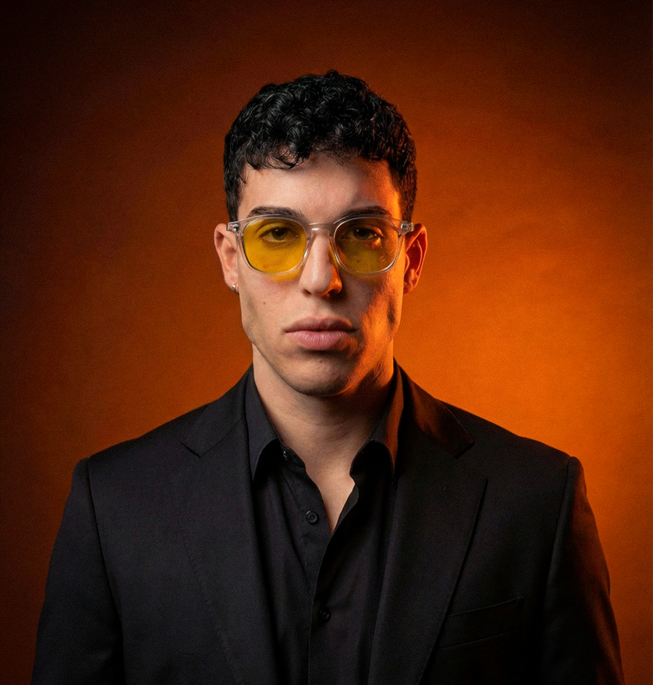 A studio portrait of a young man with dark curly hair wearing yellow-tinted glasses and a black suit.