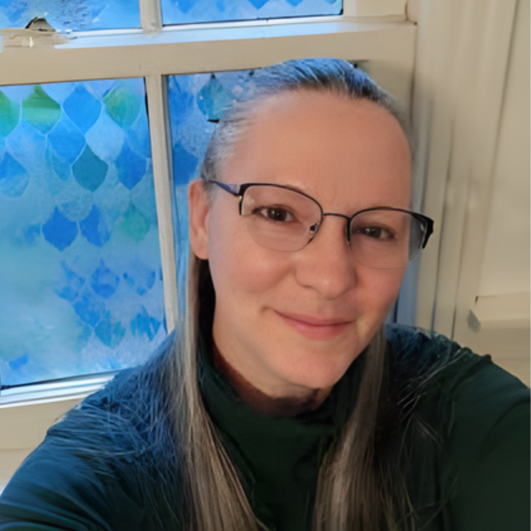 A woman with long graying hair and glasses smiles in front of a window with a blue scaled pattern.