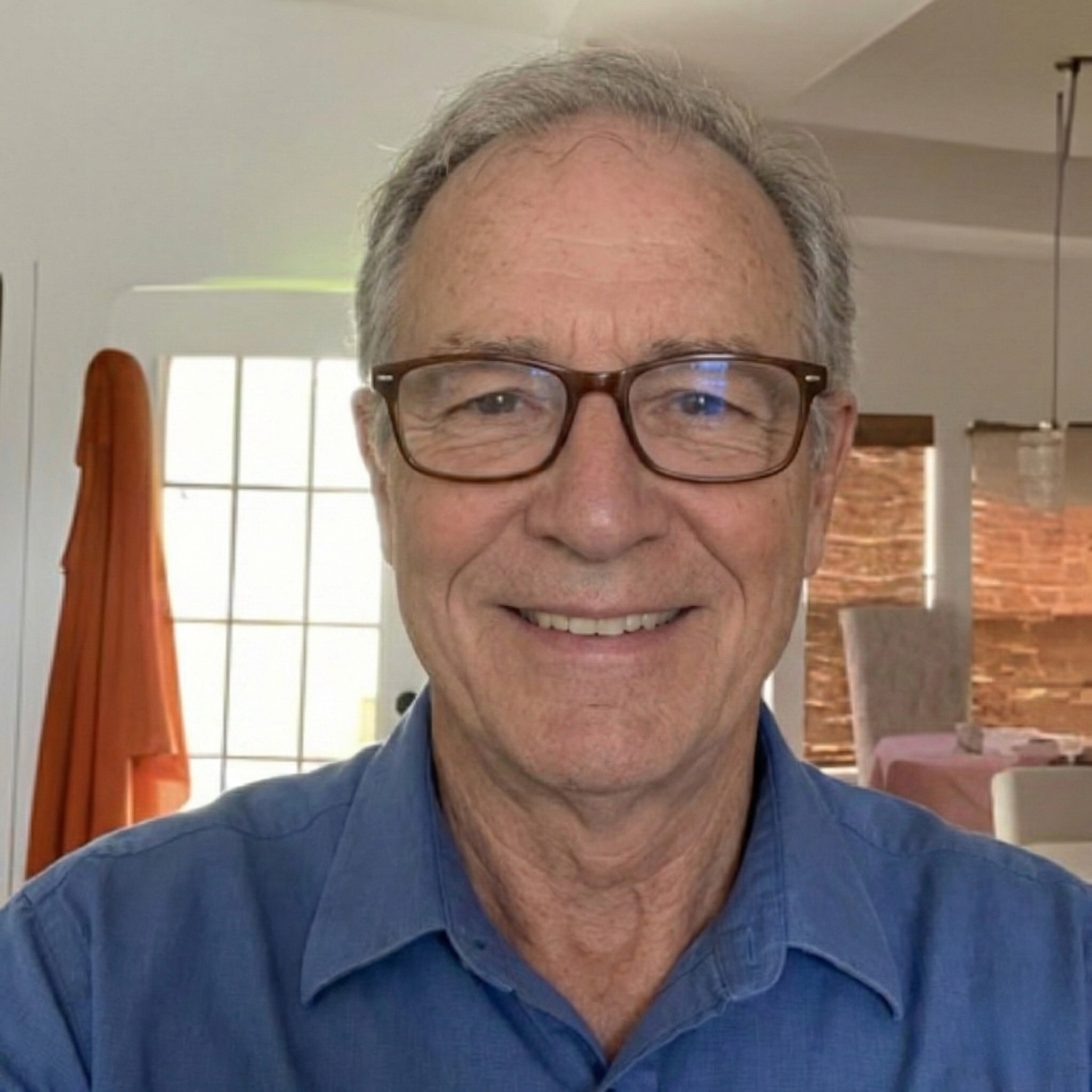 A smiling older man with gray hair and glasses, wearing a blue collared shirt, looks at the camera.