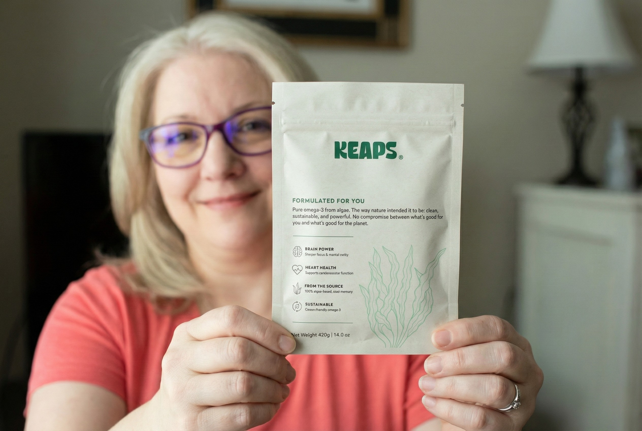 A woman with glasses holds up a white pouch of KEAPS supplements while smiling at the camera.
