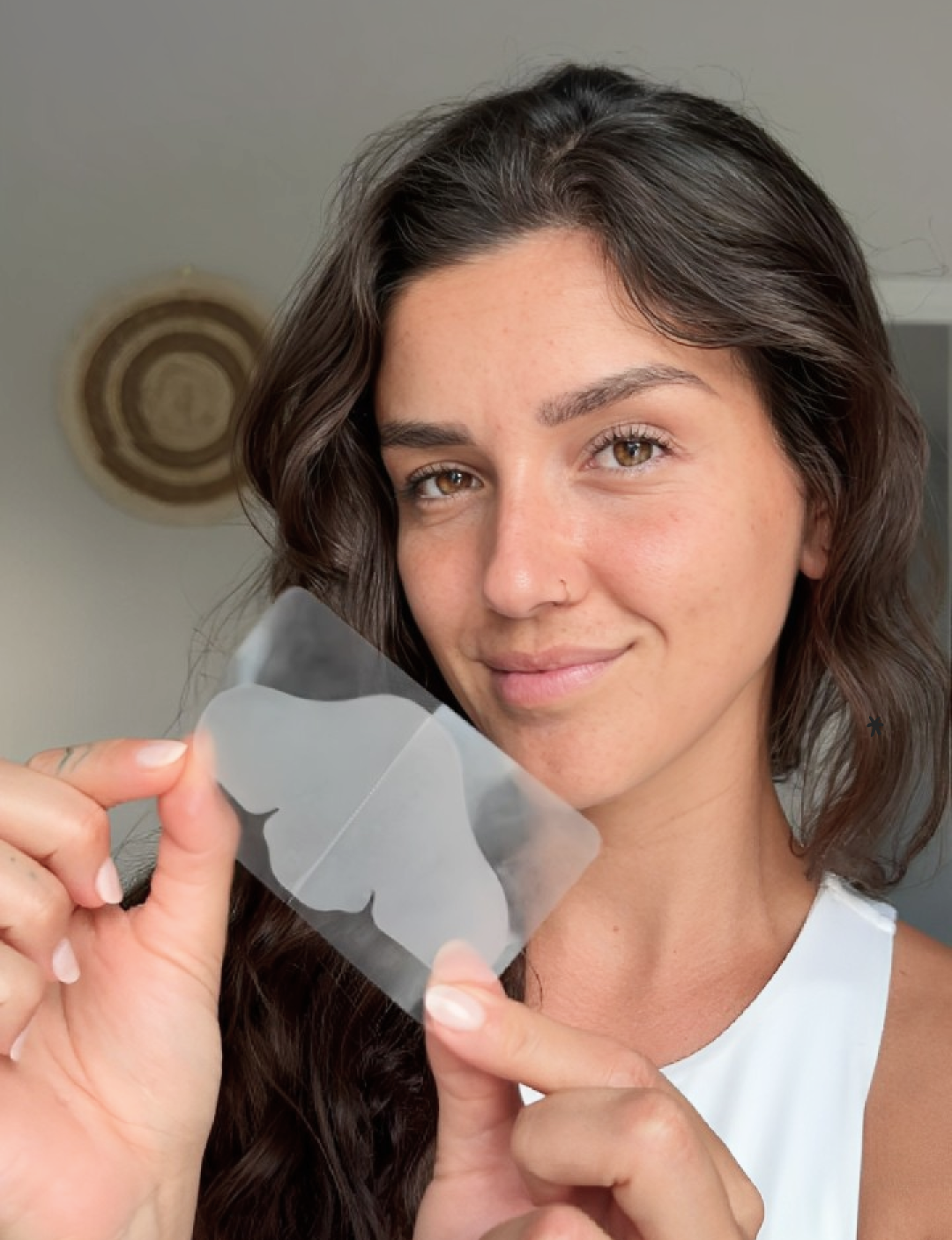 A smiling woman with dark wavy hair holds up a clear sheet with two translucent, wing-shaped patches.