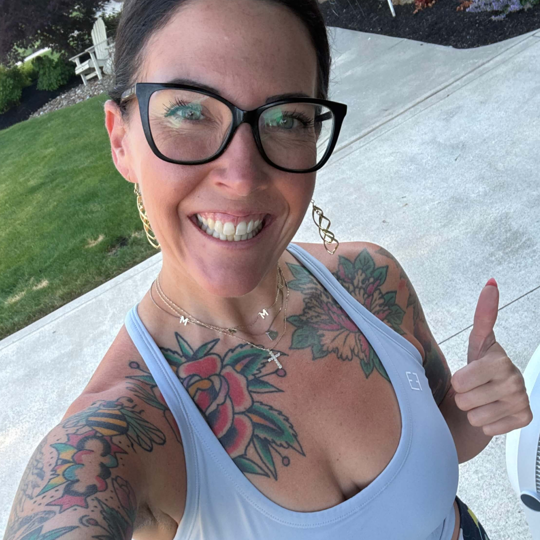 A smiling woman with glasses and tattoos gives a thumbs-up outdoors.