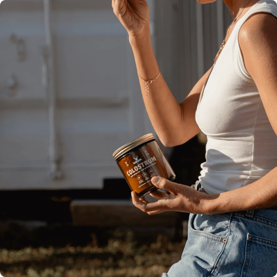 A person in a white tank top and blue jeans holds a jar of Heart & Soil Colostrum powder.