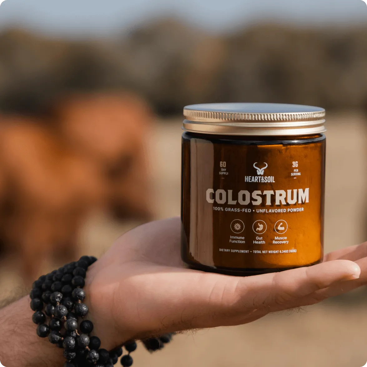 A person's hand holding a jar of Heart & Soil Colostrum dietary supplement powder outdoors.