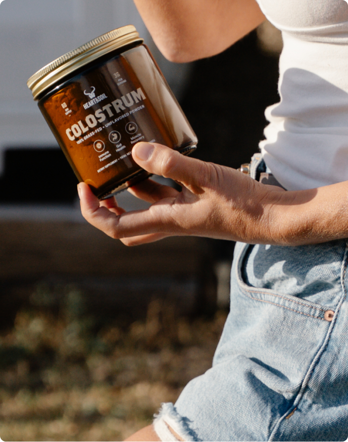 A person wearing a white shirt and denim shorts holds a jar of Heart&Soil Colostrum powder outdoors.