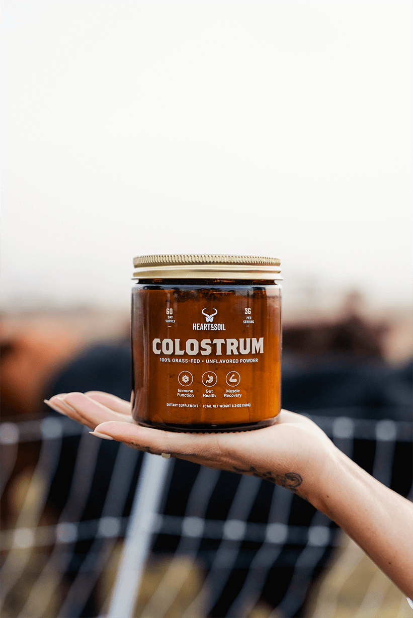 A person's hand holds a jar of Heart & Soil Colostrum powder in an outdoor setting.