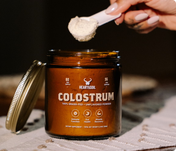 A hand holds a scoop of Heart & Soil Colostrum powder over the open amber glass jar.