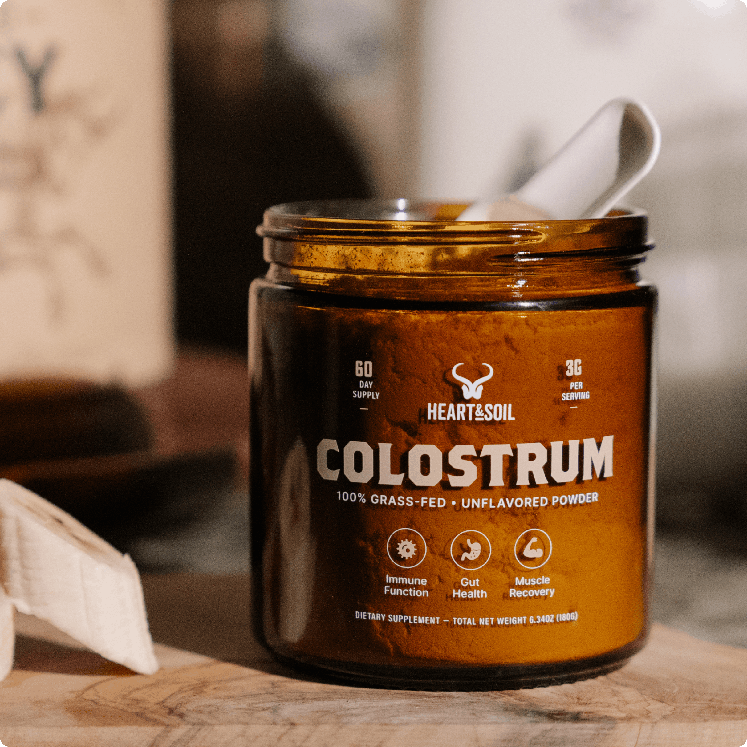 A jar of Heart & Soil Colostrum powder with a scoop, sitting on a wooden surface.