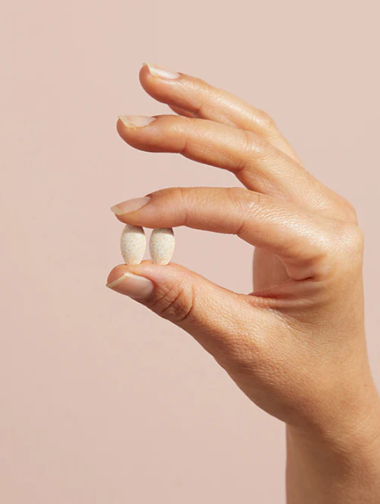 A hand holding two white pills against a beige background.