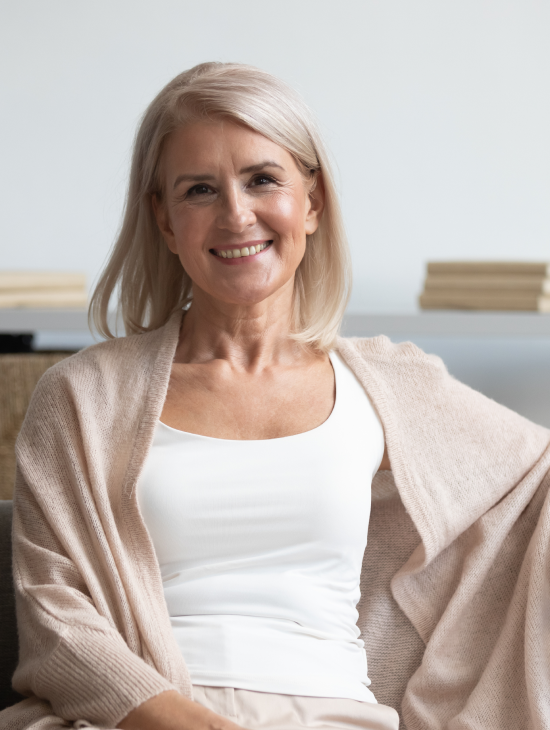 Smiling person with light hair wearing a white top and beige cardigan, sitting indoors.