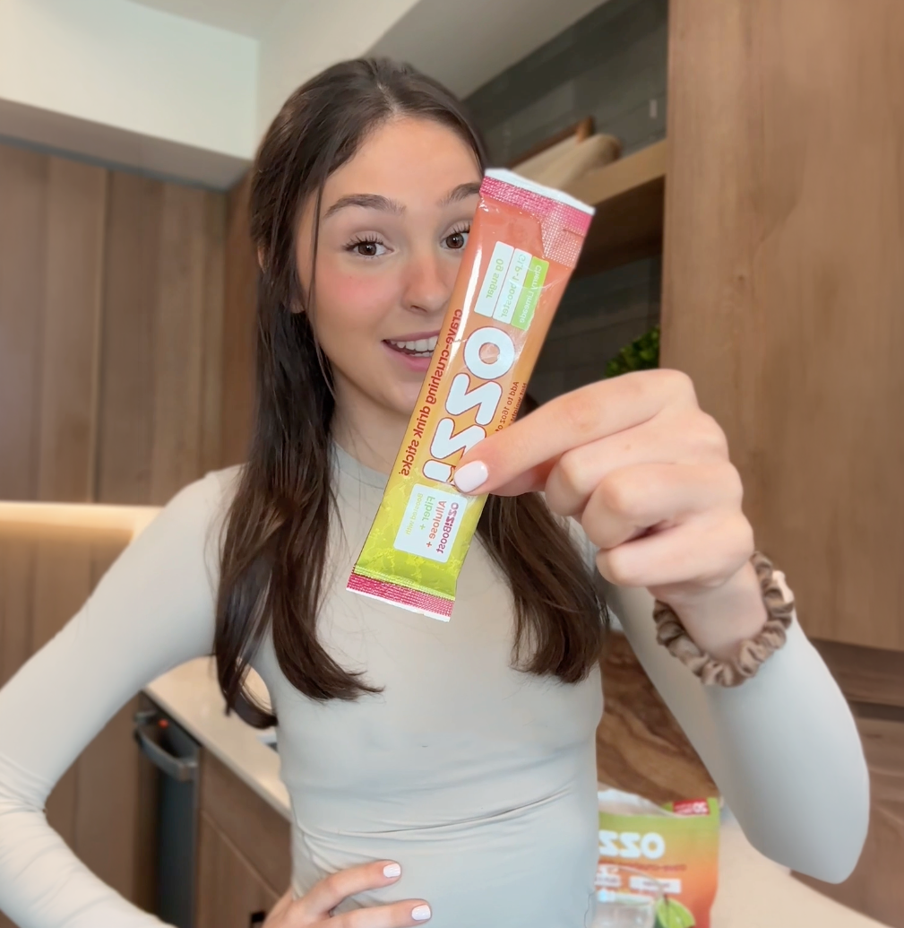 A young woman smiles while holding an OSSO drink mix packet towards the camera in a kitchen.