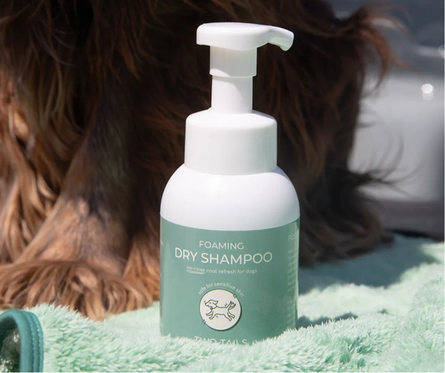 A bottle of foaming dry shampoo for dogs sits on a green towel in front of a brown dog.