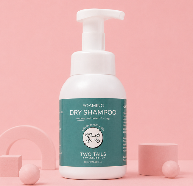 A bottle of Two Tails Pet Company Foaming Dry Shampoo for dogs against a pink background with geometric shapes.