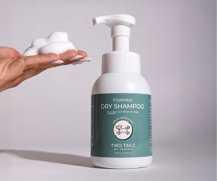 A hand holds white foam next to a bottle of Two Tails Pet Company foaming dry shampoo.