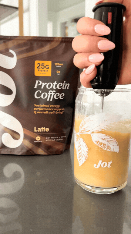 Hand using a frother in a glass of protein coffee next to a coffee bag.