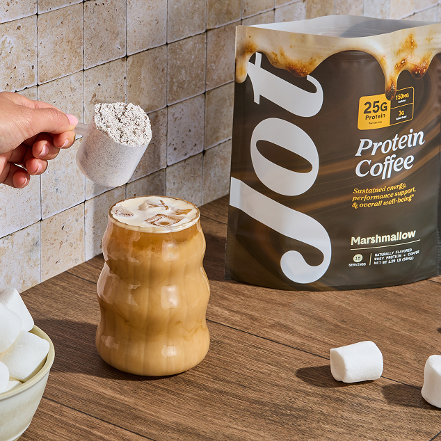 A hand scoops protein coffee powder into an iced coffee, next to the product bag and marshmallows.
