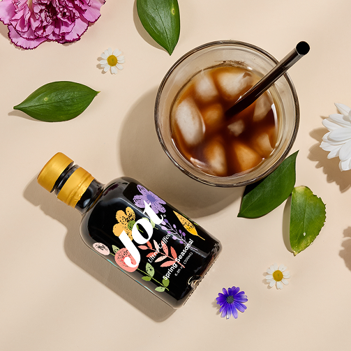 Bottle of Jot coffee next to iced coffee glass, surrounded by flowers and leaves.