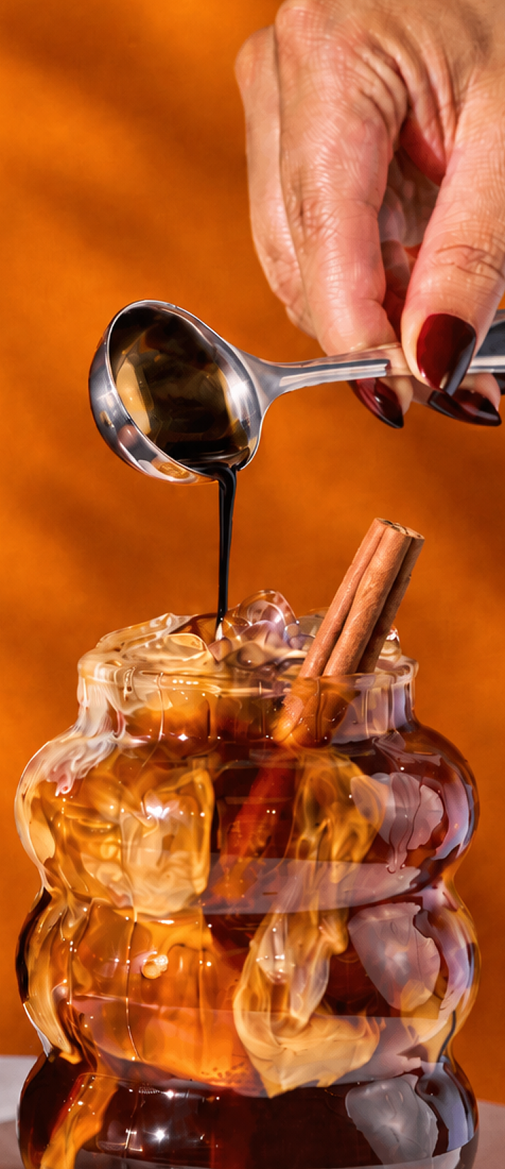 A hand pours dark syrup from a spoon into an iced drink with cinnamon sticks.