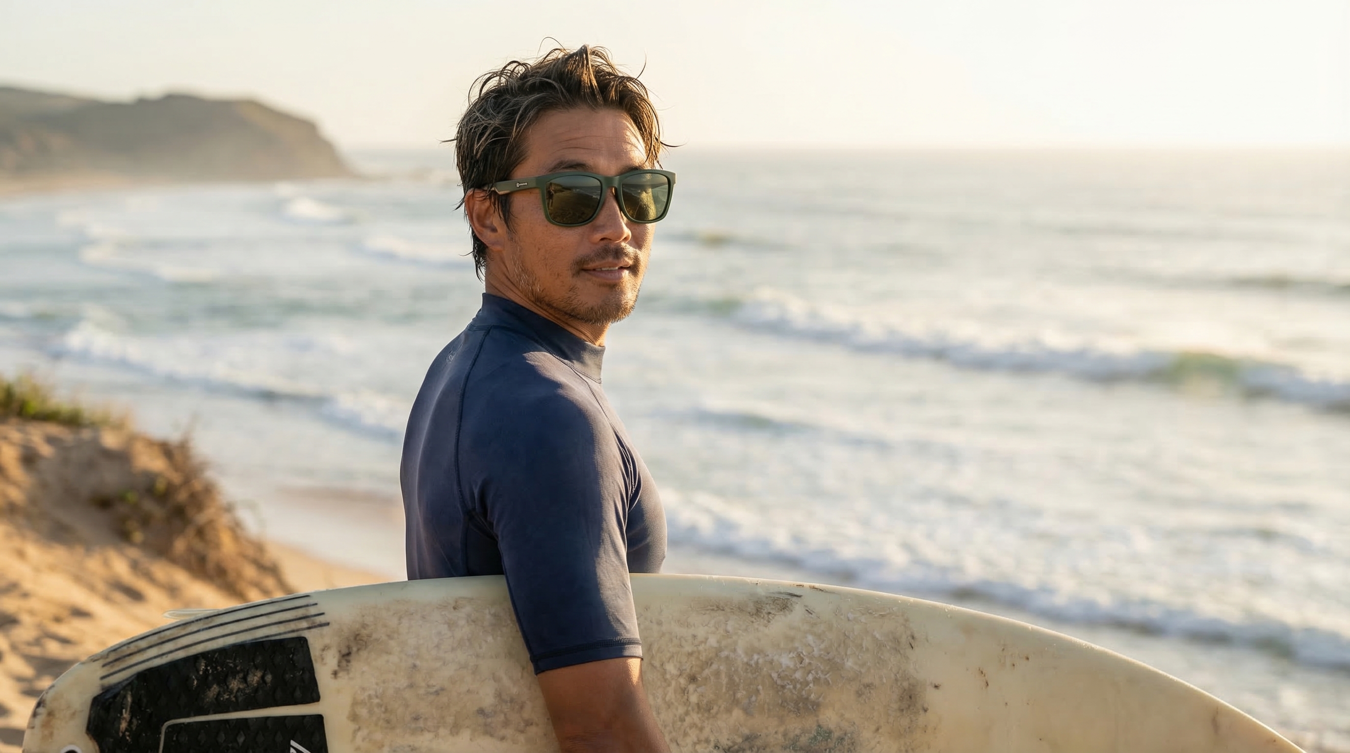 A man in sunglasses and a rash guard holds a surfboard at the beach, looking back.