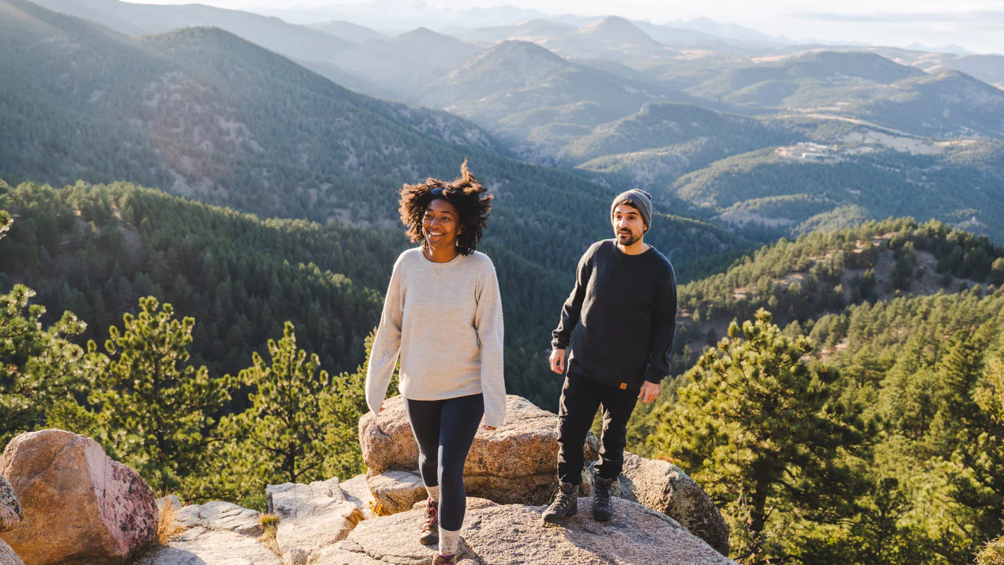 Two people hiking on a rocky mountain with forested hills in the background.
