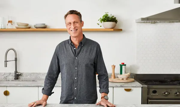 A smiling man in a grey button-down shirt stands behind a counter in a modern kitchen.