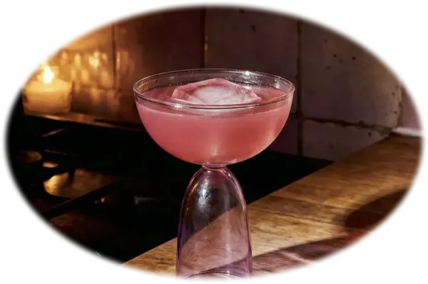 A Magic Calm with a large ice cube in a unique, bell-shaped glass sits on a wooden counter, illuminated by the warm glow of nearby lit candles.