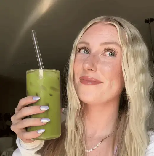 A woman with blonde hair smiles while holding a green smoothie in a clear glass.