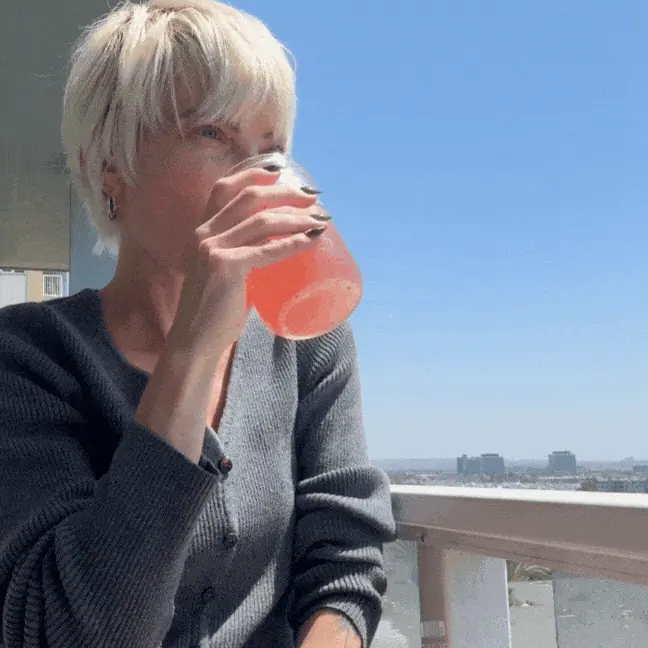 A person with short blonde hair sips a pink drink from a glass on a sunny balcony.