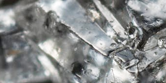 A close-up, macro photograph of a silvery, crystalline metal with a rough, textured surface.