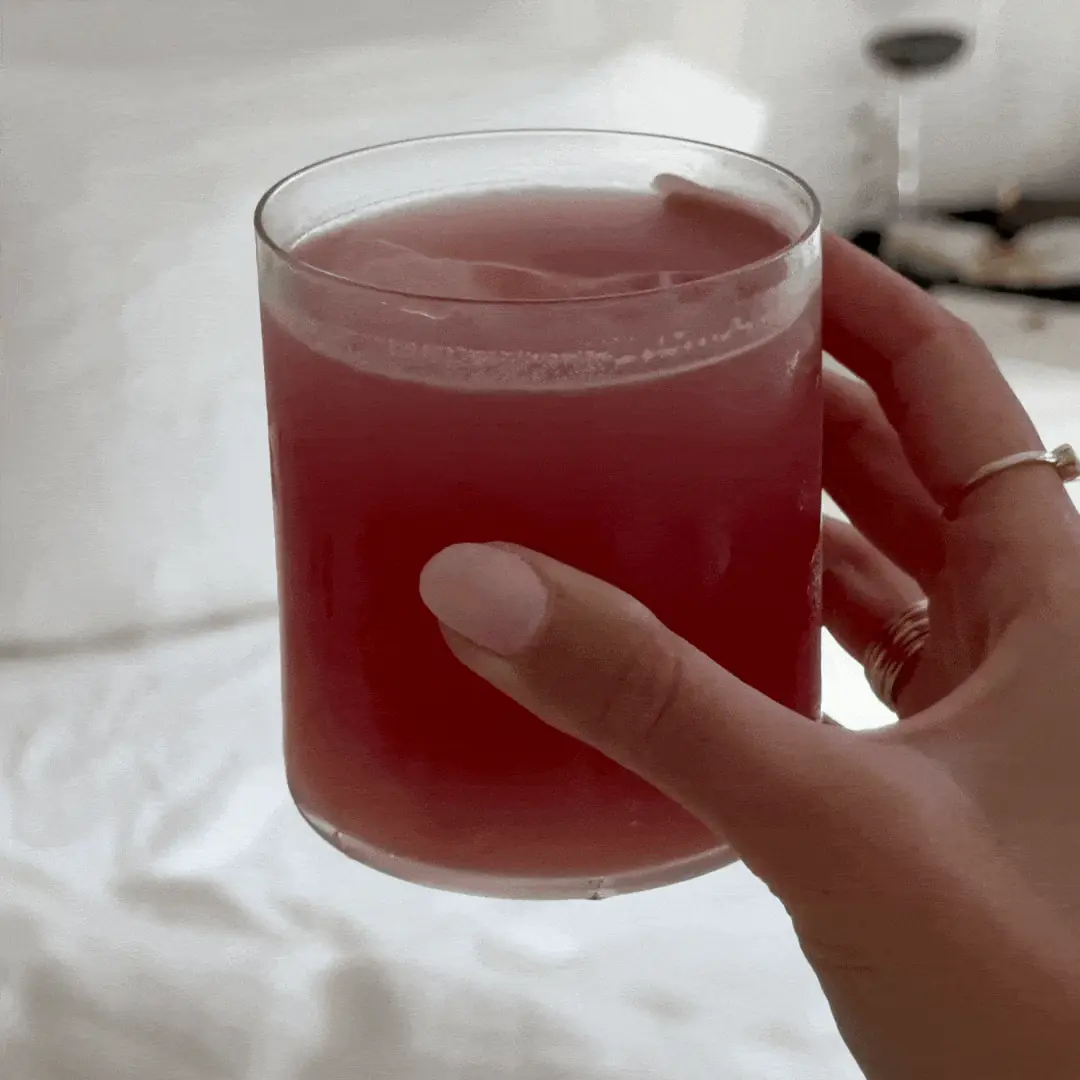 A hand with light pink nails holds a glass filled with a reddish-pink liquid.
