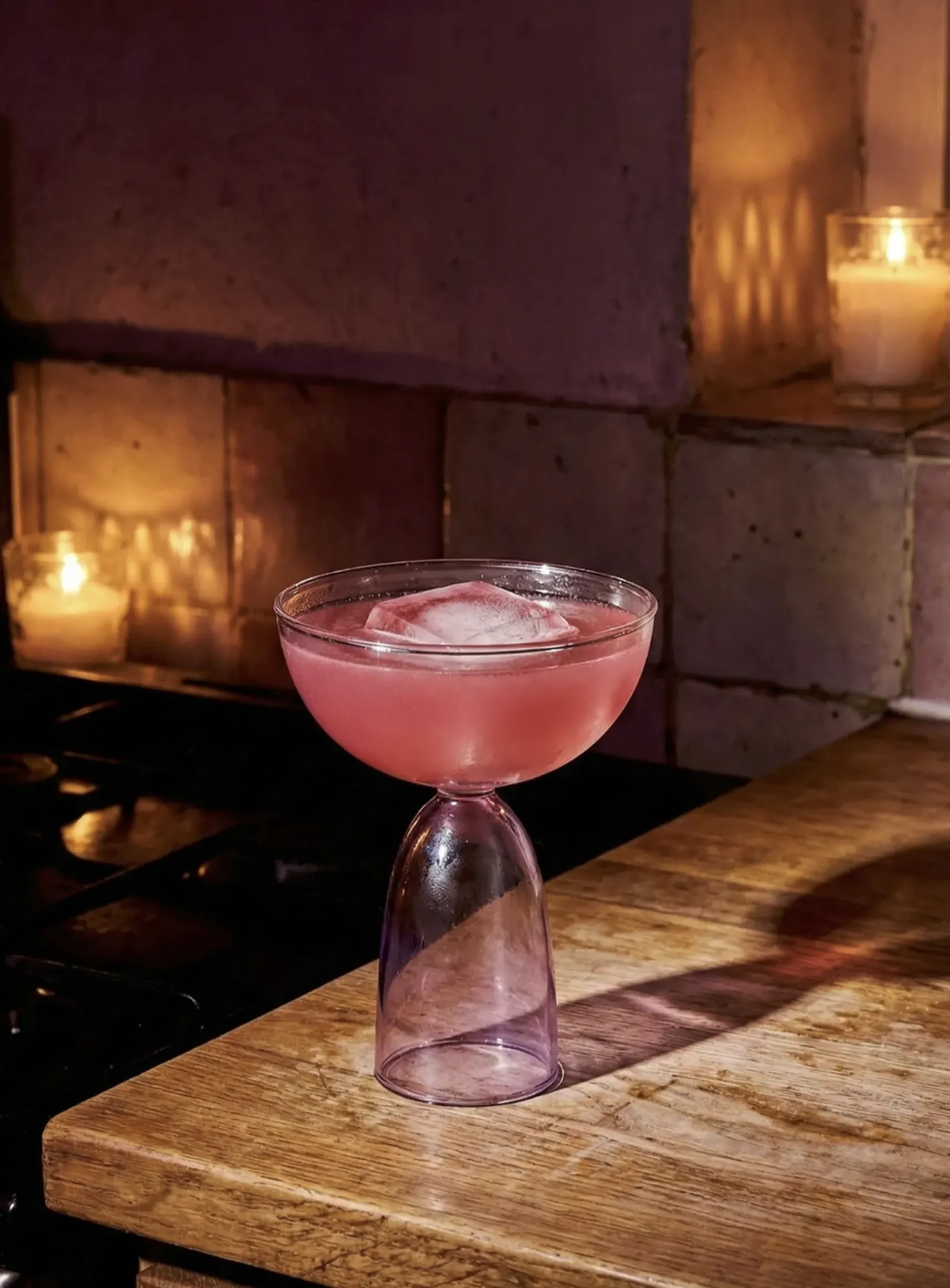 A pink cocktail in a coupe glass sits on a wooden counter, illuminated by candlelight.