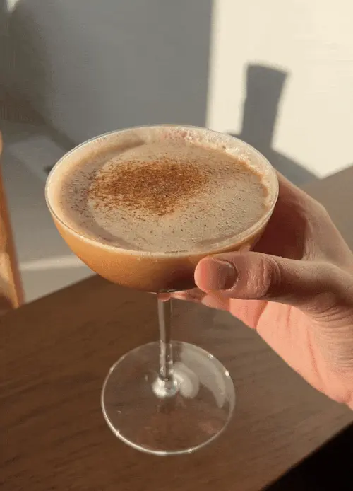Hand holding a creamy cappuccino in a coupe glass, dusted with cinnamon, glowing in warm sunlight above a wooden table.