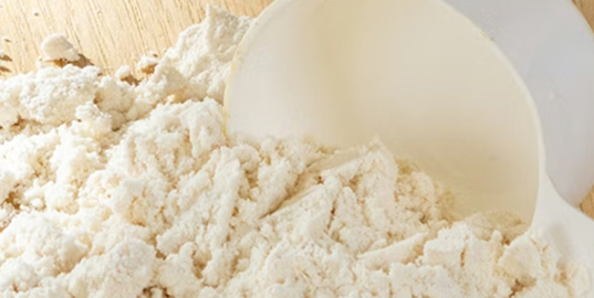 A close-up of a white plastic scoop resting in a pile of off-white powder on a wooden surface.