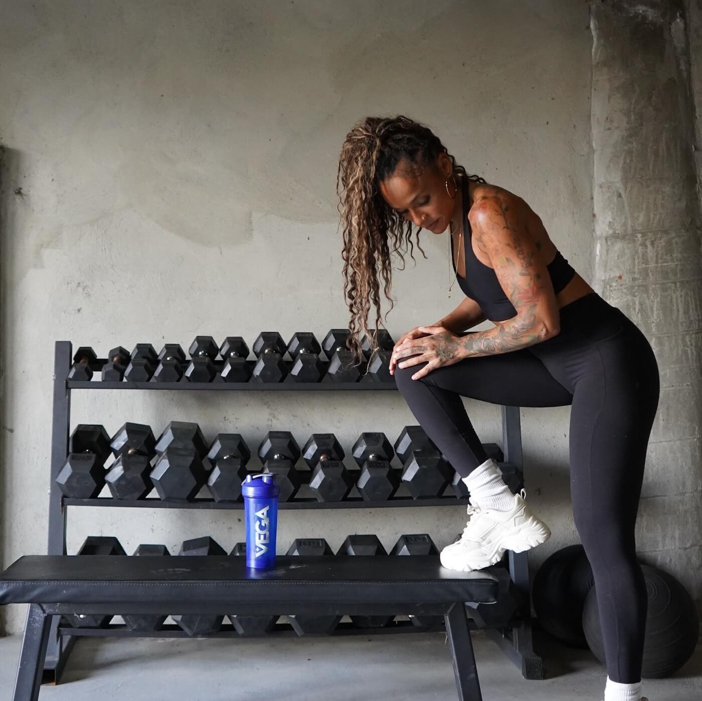 Person in workout attire stretching near a rack of dumbbells and a bench.