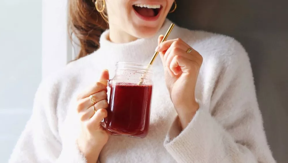 A woman in a white sweater smiles while holding a glass mug of red juice with a straw.