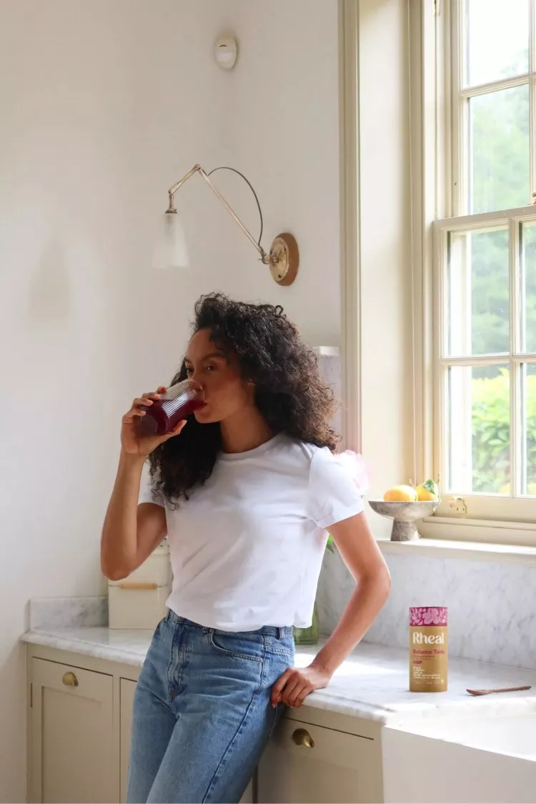 A woman with curly hair stands in a kitchen drinking a red beverage from a glass.