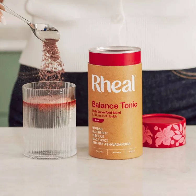 A person spoons Rheal Balance Tonic powder into a glass of water next to the product container.