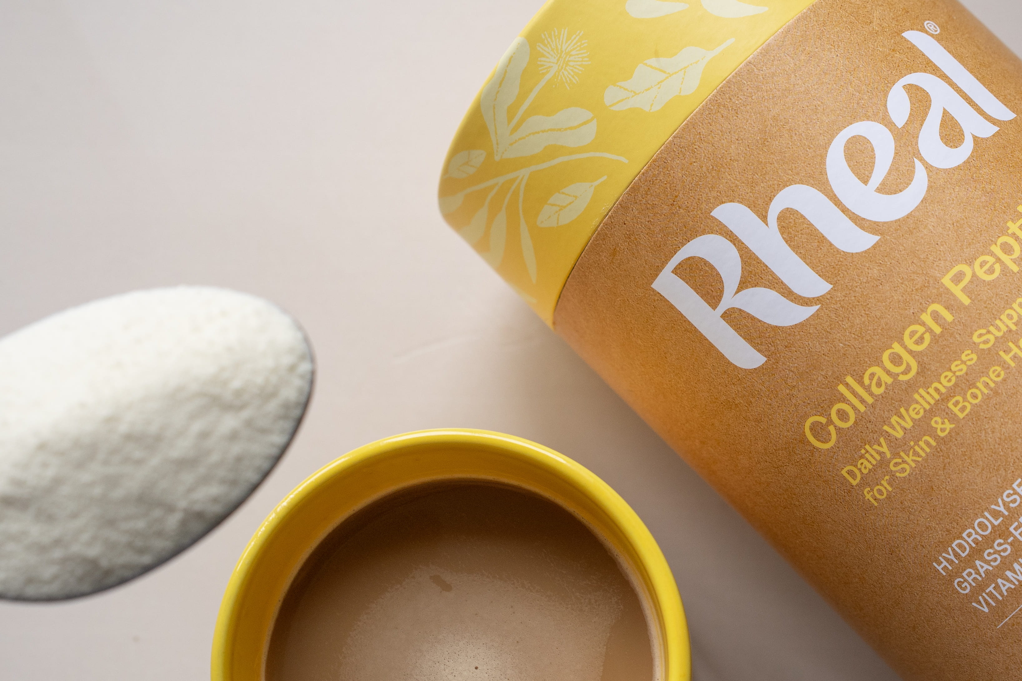 A flat lay of a Rheal Collagen Peptides container, a yellow mug, and a spoon of powder.