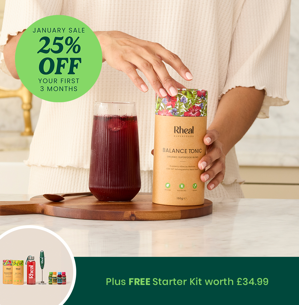 Person holding a Rheal Balance Tonic container next to a glass of red juice.