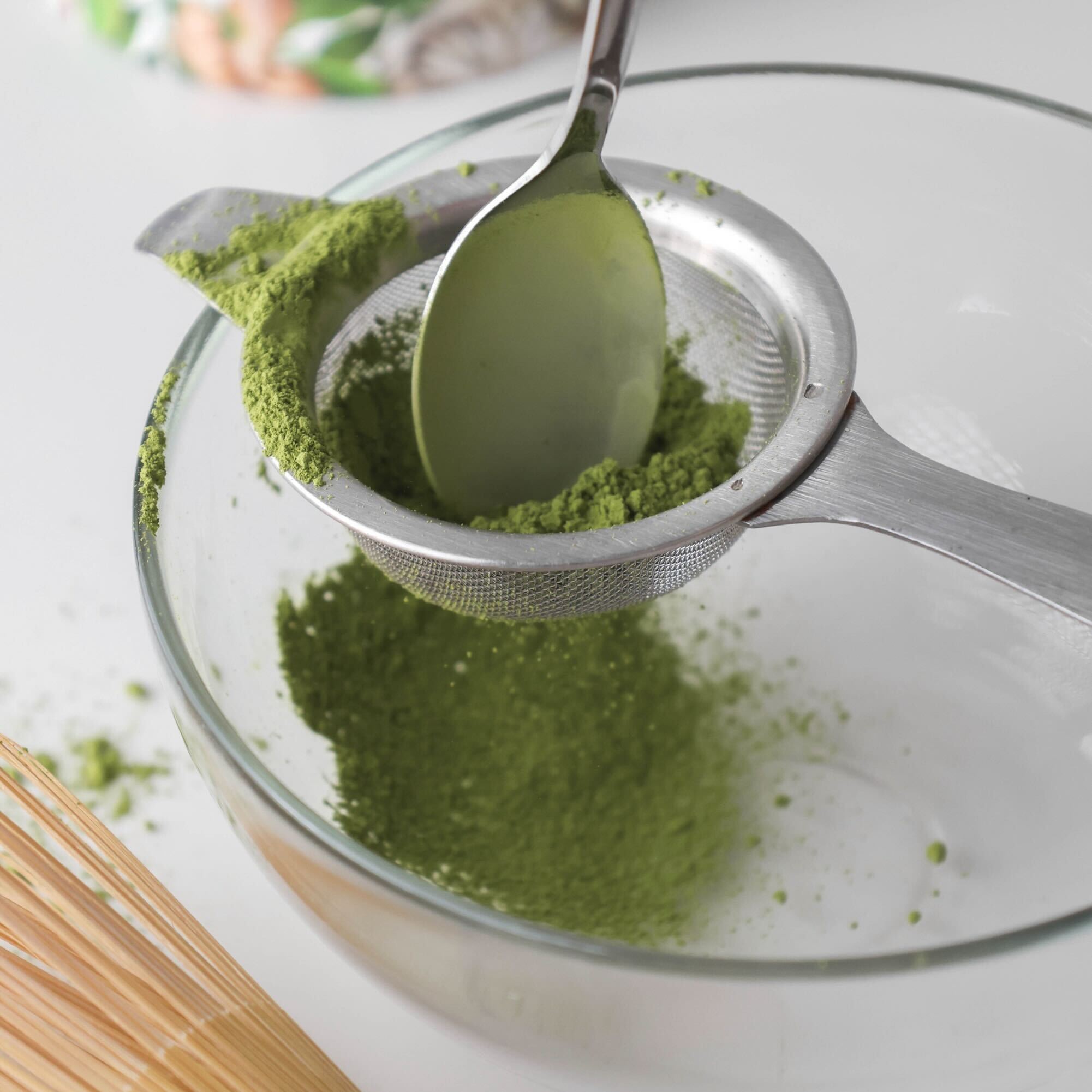 Green powder being sifted into a glass bowl with a spoon and strainer.