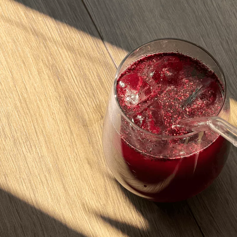 A glass of a red, pulpy beverage with ice and a glass straw on a wooden surface.